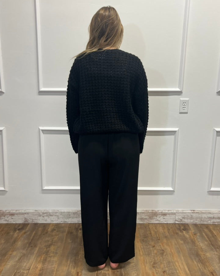 A woman stands with her back to the camera wearing a black textured knit sweater and black wide-leg pants in front of a white paneled wall on a wood floor.