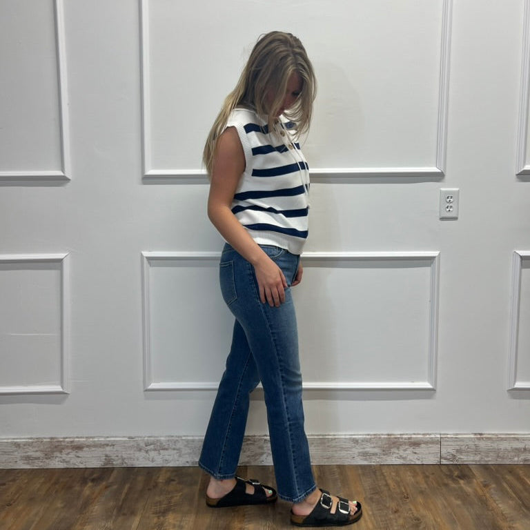 A woman stands in profile wearing a sleeveless navy and white striped top, blue jeans, and black slide sandals, standing against a white paneled wall on a wood floor.