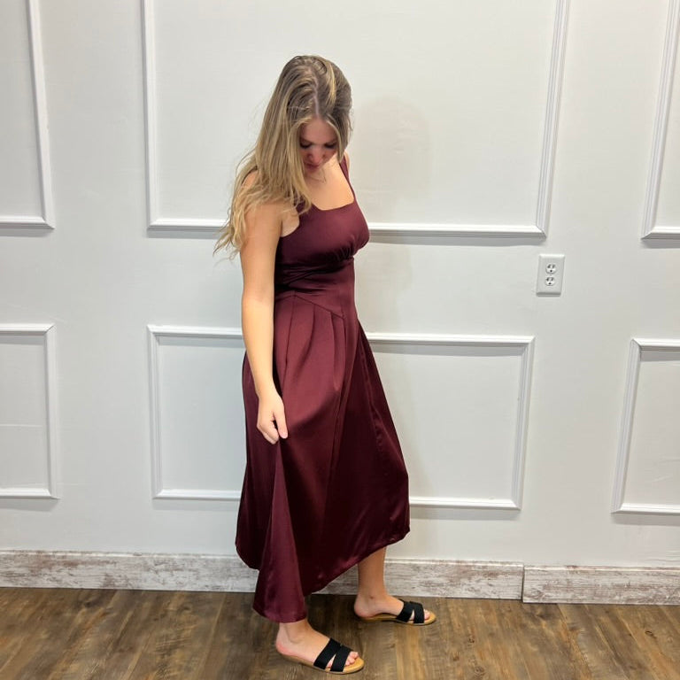 A woman modeling a sleeveless, floor-length satin-like dress in deep burgundy, standing on a wood floor against a white paneled wall.