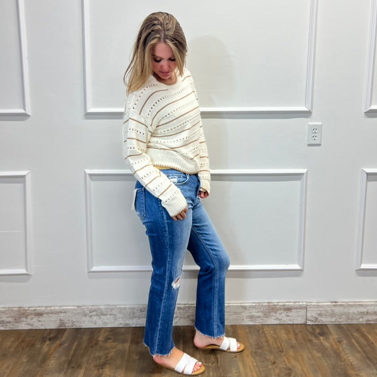 Woman wearing a cream sweater and blue jeans standing against a white paneled wall.