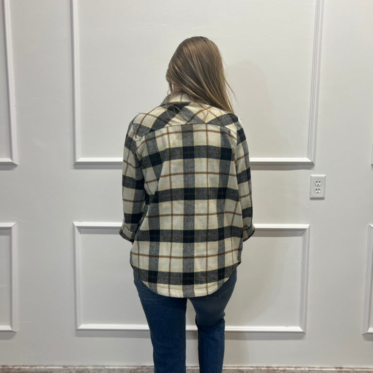 A woman viewed from behind wearing a black and cream plaid flannel shirt and dark blue jeans, standing in front of a white paneled wall.