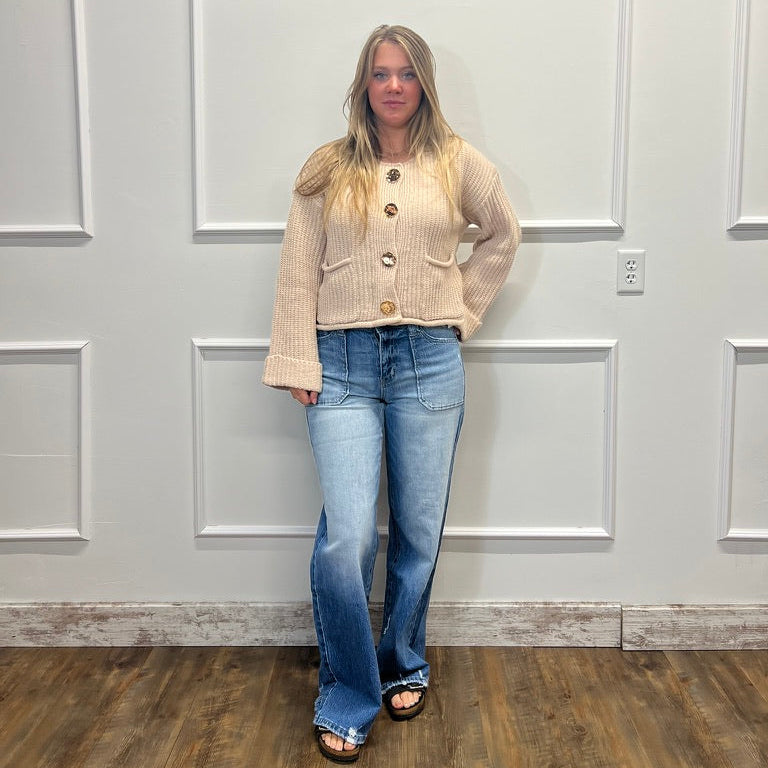 A woman stands in a room with white paneled walls and a wood floor, wearing a beige button-front cardigan with wide sleeves and blue flare jeans.
