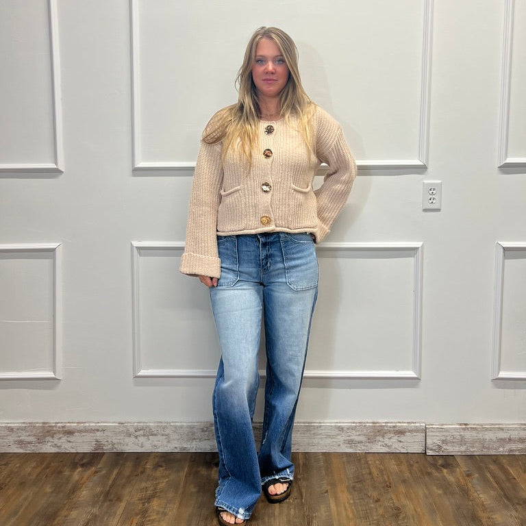 A woman stands in a room with white paneled walls and a wood floor, wearing a beige button-front cardigan with wide sleeves and blue flare jeans.
