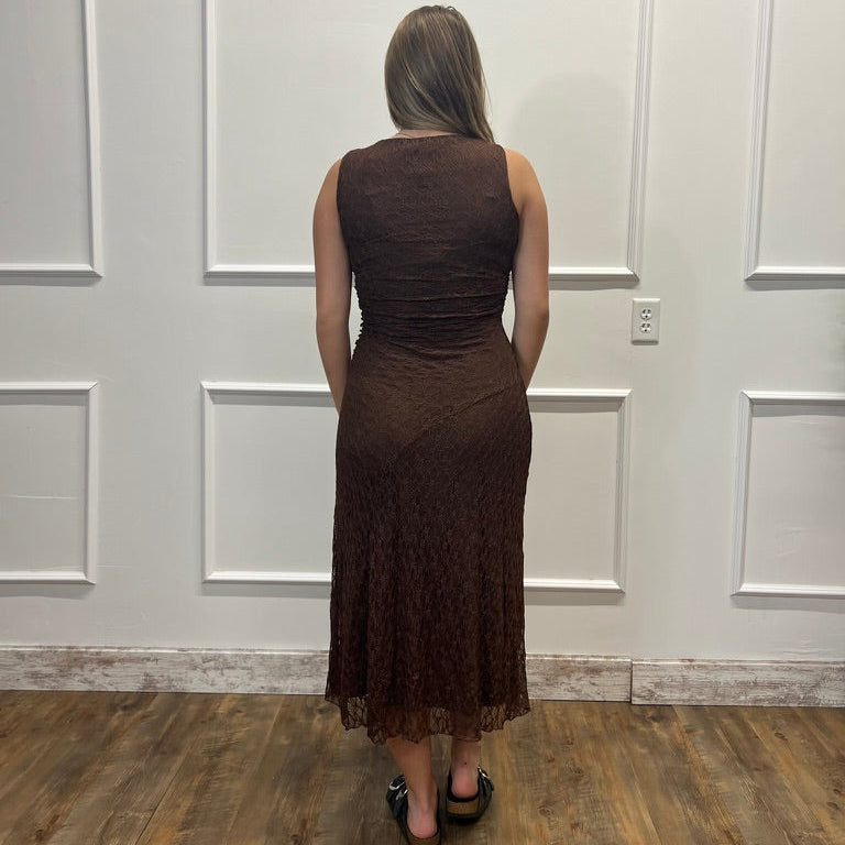 A woman viewed from behind wearing a sleeveless, knee-length brown lace dress, standing on a wood floor against a white paneled wall.