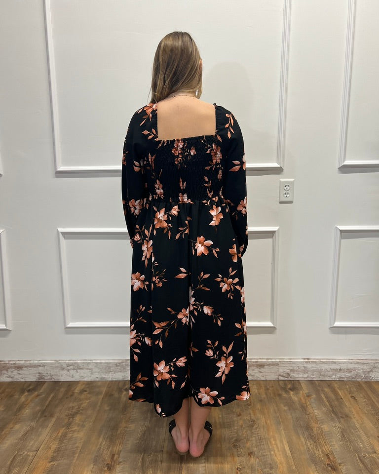 A woman viewed from behind wearing a black long-sleeve maxi dress with a pink and peach floral pattern, standing on a wood floor against a white paneled wall.