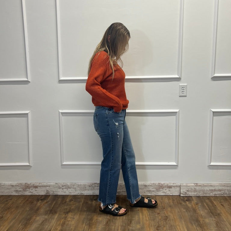A woman stands in profile wearing a rust-orange long-sleeve knit top, blue jeans, and black sandals against a white paneled wall with a wood floor.