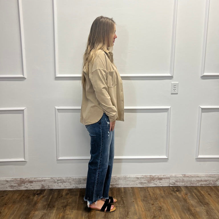 A woman stands in profile wearing a beige button-up shirt and blue jeans, standing on a wood floor against a white paneled wall.