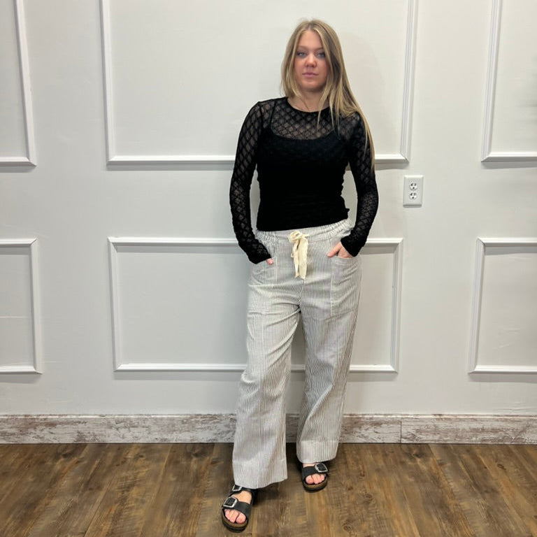 A woman stands in a room with white paneled walls and a wood floor, wearing a black long-sleeve mesh top, light beige wide-leg pants, and black sandals.