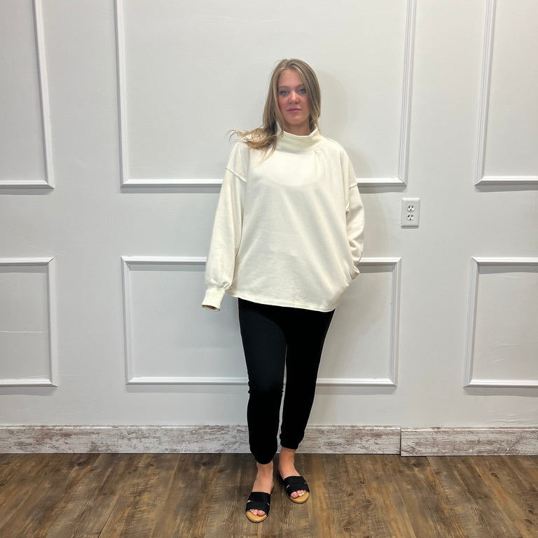 A woman stands in a room with white paneled walls and a wood floor, wearing an oversized cream mock-neck long-sleeve top, black pants, and black slide sandals.