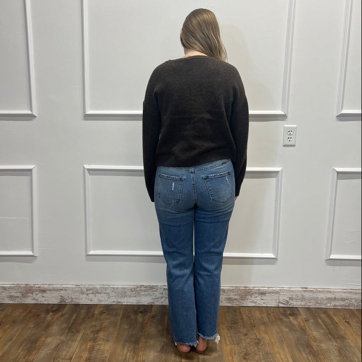 A woman viewed from behind wearing a dark charcoal sweater and blue jeans, standing on a wood floor against a white paneled wall.
