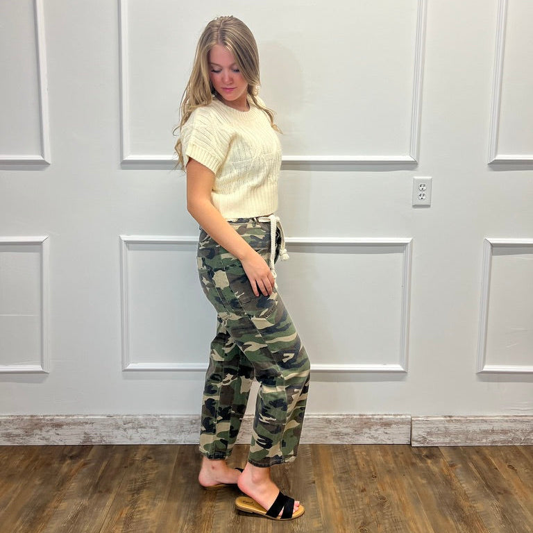 A woman stands in a room with white paneled walls and a wood floor, wearing a cream short-sleeve knit top and camouflage drawstring pants with black slide sandals.