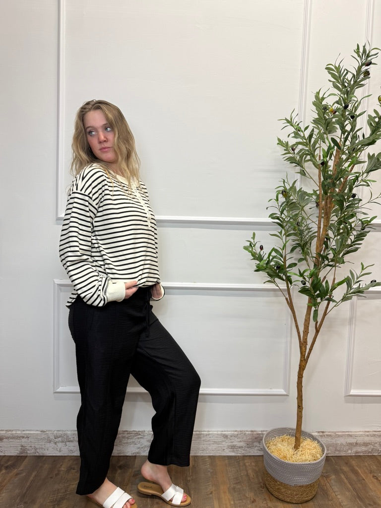 Woman in a striped shirt and black pants leaning against a white wall with a plant next to her.