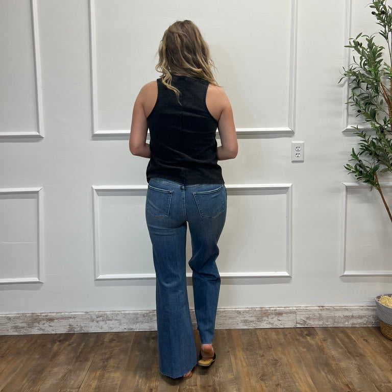 Person wearing a black sleeveless top and blue jeans standing in a room with white paneled walls and wooden flooring.