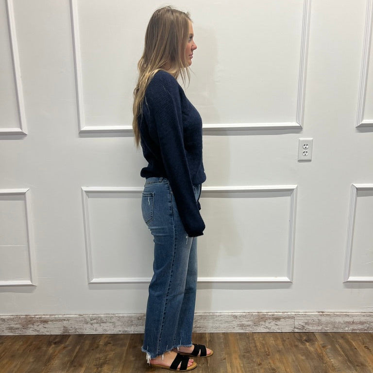 A woman stands in profile wearing a dark navy long-sleeve knit top and blue flare jeans with black slide sandals, standing on a wood-look floor against a white paneled wall.