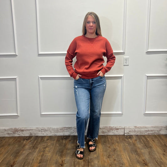 A woman stands in a room with white paneled walls and a wood floor, wearing a rust-orange crewneck sweater, blue jeans, and black sandals.