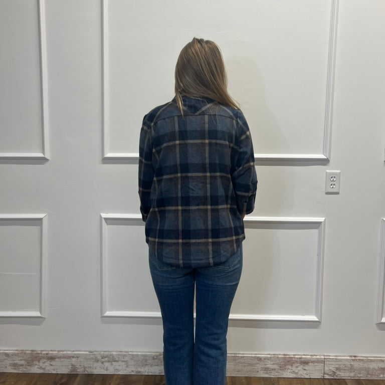 A woman viewed from behind wearing a dark navy and black plaid flannel shirt and blue jeans, standing in front of a white paneled wall.