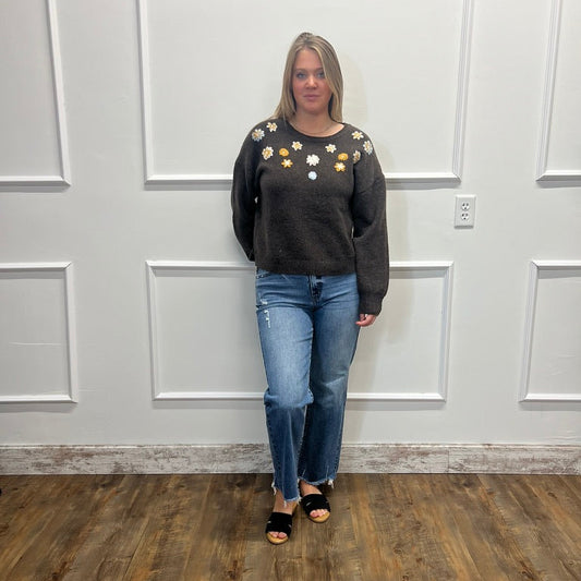 A woman stands in a room with white paneled walls and a wood floor, wearing a dark charcoal gray crewneck sweater with white and yellow flower embellishments on the chest, paired with blue jeans and black slide sandals.
