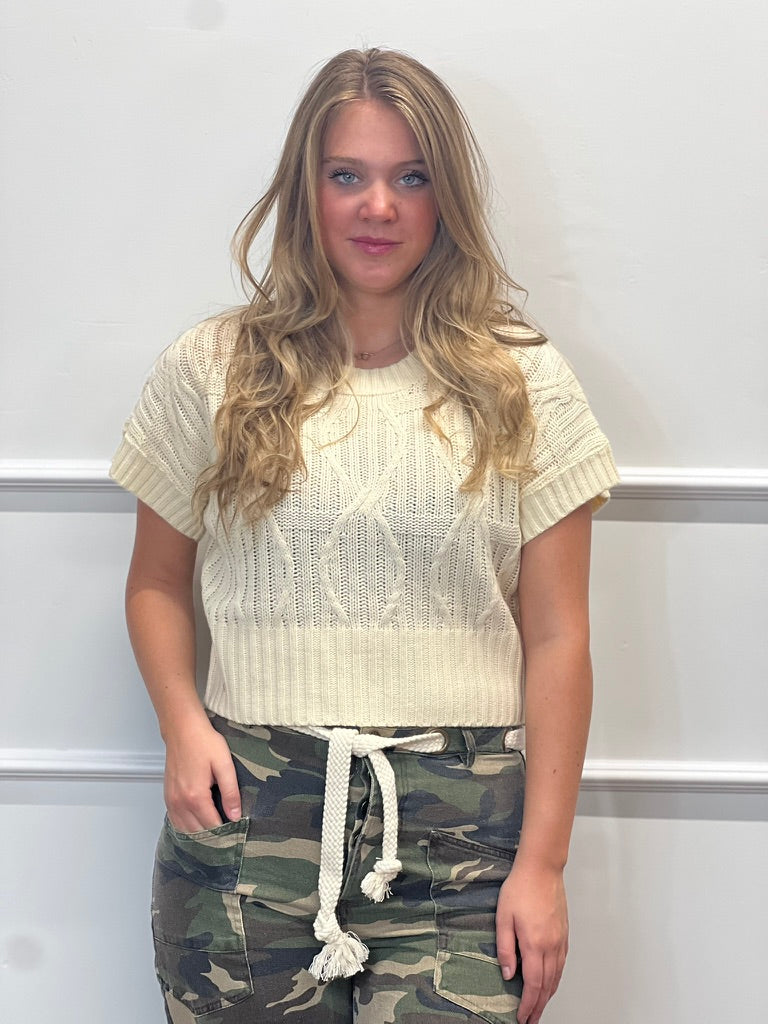 A woman with long blonde hair stands against a white paneled wall, wearing a cream-colored short-sleeve cable-knit cropped sweater and camouflage drawstring pants.