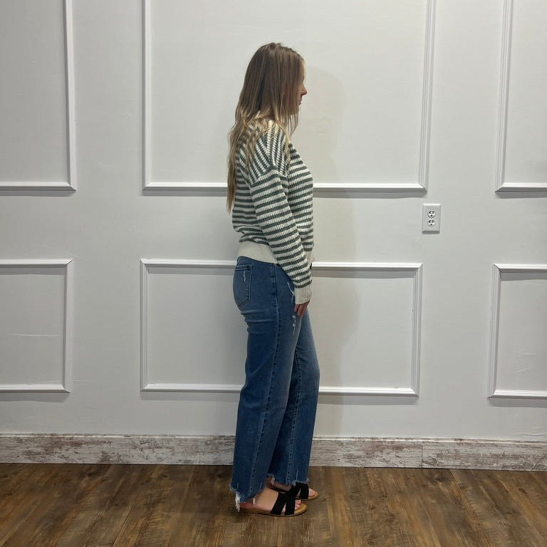 A woman stands in profile against a white paneled wall on a wood floor, wearing a green and white striped long-sleeve top, blue jeans, and black sandals.