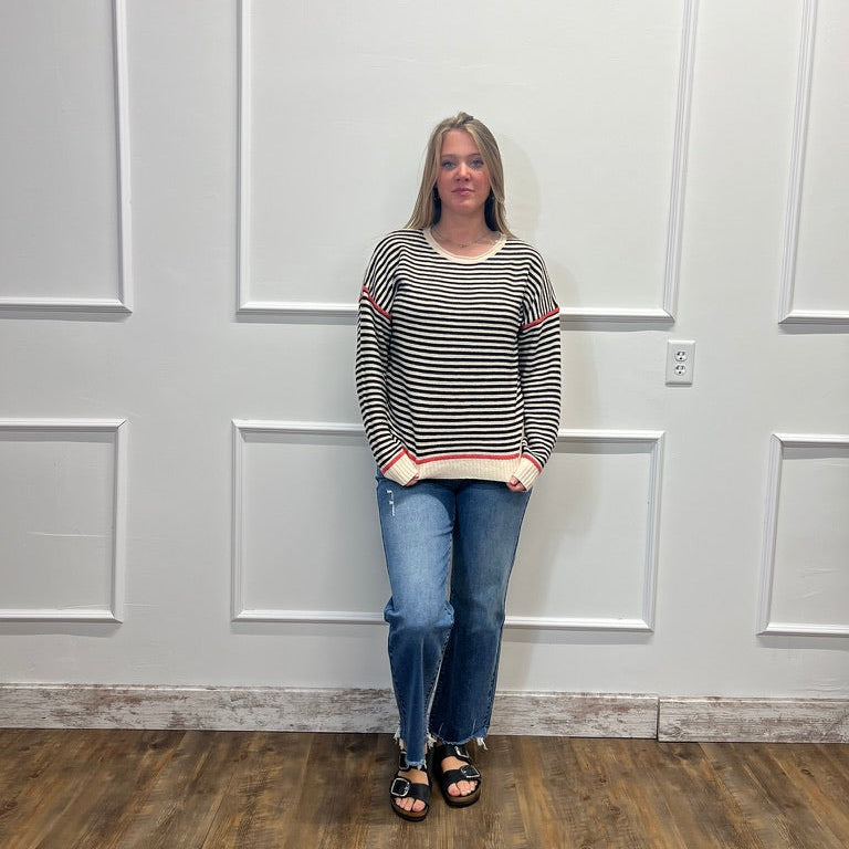 A woman stands in a room with white paneled walls and a wood floor, wearing a black and white striped long-sleeve top with red accents, blue jeans, and black sandals.