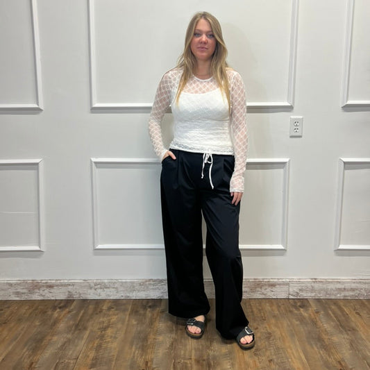 A woman stands in a room with white paneled walls and a wood floor, wearing a white long-sleeve top with a sheer patterned overlay and black wide-leg drawstring pants.