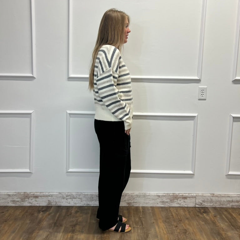 A woman stands in profile wearing a cream knit sweater with thin dark gray horizontal stripes and black wide-leg pants, standing on a wood floor against a white paneled wall.