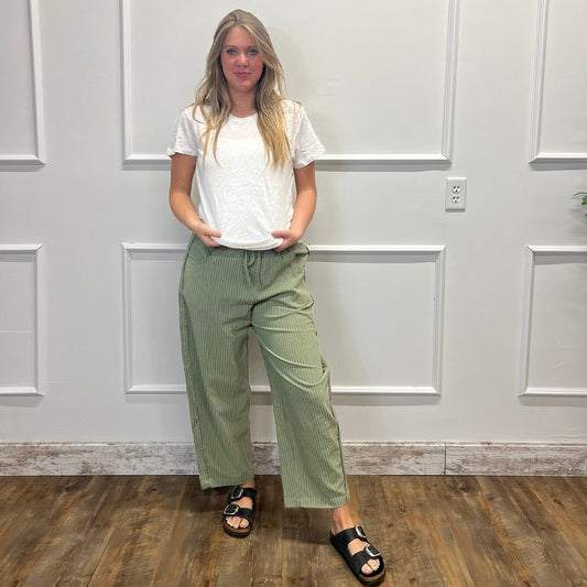 A woman stands against a white paneled wall wearing a white short-sleeve t-shirt, sage green wide-leg cropped pants, and black slide sandals.