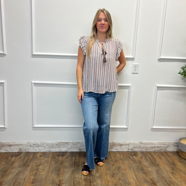 A woman stands in a room with white paneled walls and wood-look flooring, wearing a sleeveless striped top with ruffled shoulders, blue flare jeans, and black slide sandals.