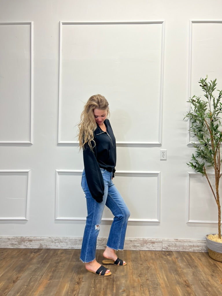 Woman in black top and blue jeans standing in a room with white paneled walls and a plant.