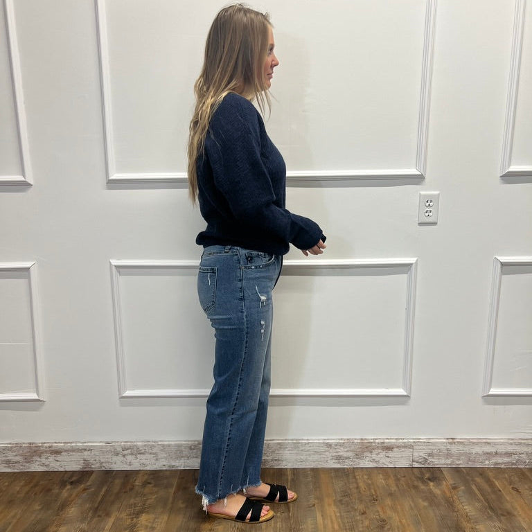 A woman stands in profile wearing a dark navy long-sleeve knit sweater and blue distressed flare jeans with black slide sandals, set against a white paneled wall and wood-look floor.