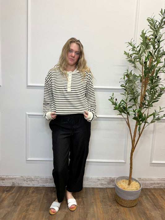 Person wearing a striped shirt and black pants standing in a room with a plant.