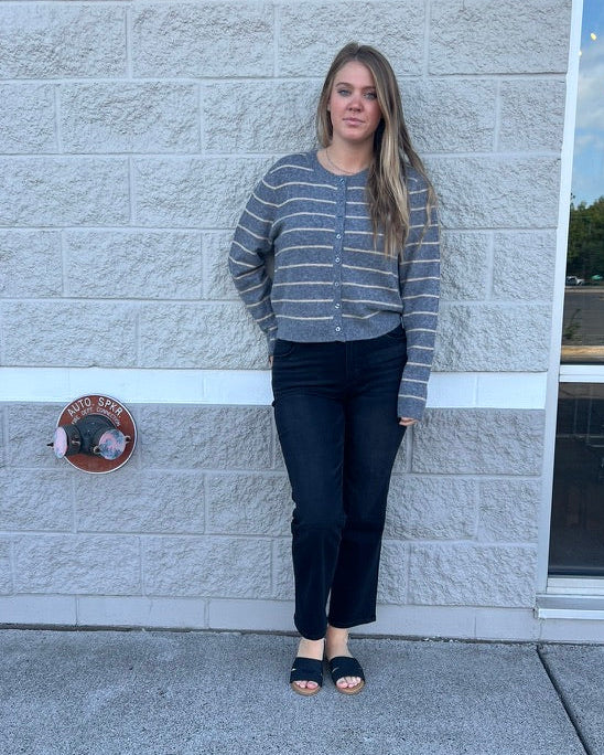 A woman stands outdoors against a light gray stone wall wearing a gray and white striped knit cardigan, dark jeans, and black slide sandals.