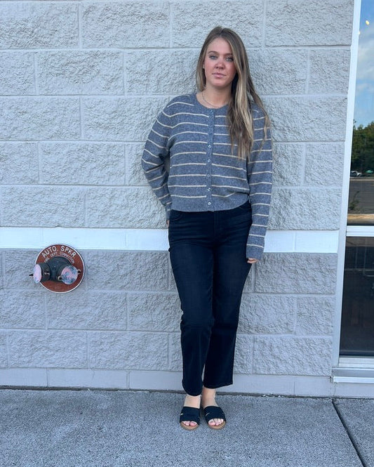 A woman stands outdoors against a light gray stone wall wearing a gray and white striped knit cardigan, dark jeans, and black slide sandals.