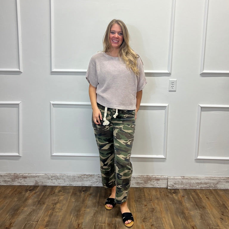 A woman stands in a room with white paneled walls and a wood floor, wearing a light gray short-sleeve top, camouflage pants, and black slide sandals.