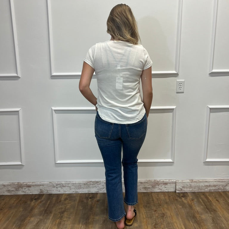 A woman viewed from behind wearing a white short-sleeve t-shirt and blue jeans, standing on a wood floor against a white paneled wall.