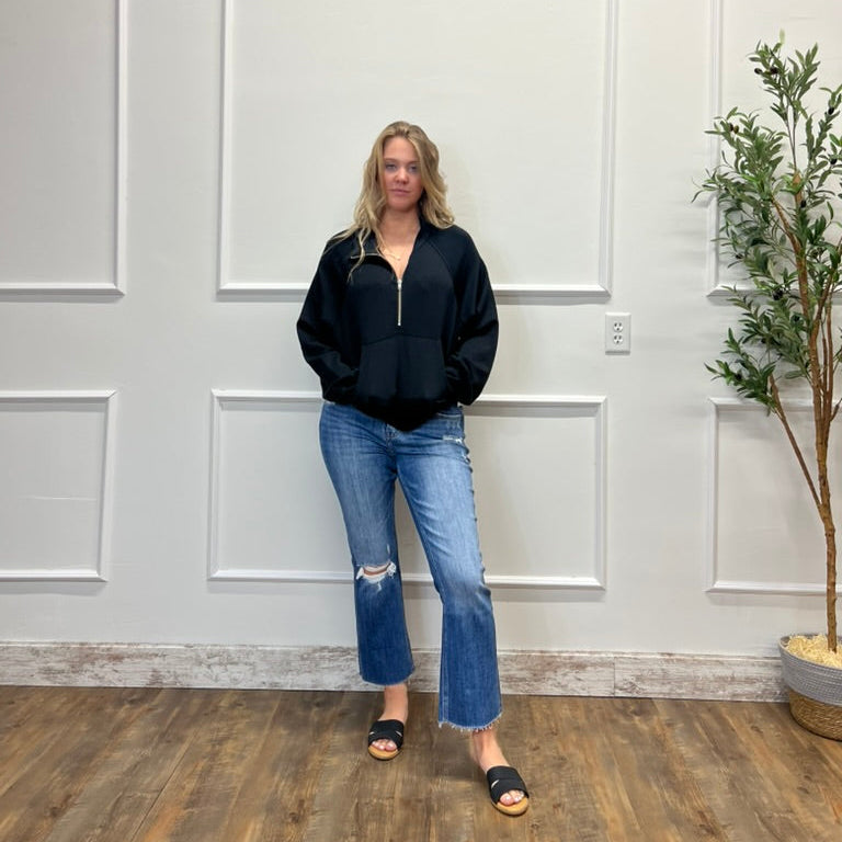 Woman wearing a black jacket and blue jeans standing in a room with white paneled walls and a plant.