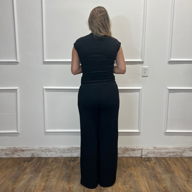 A woman viewed from behind wearing a black sleeveless jumpsuit with wide legs, standing in front of a white paneled wall on a wood floor.