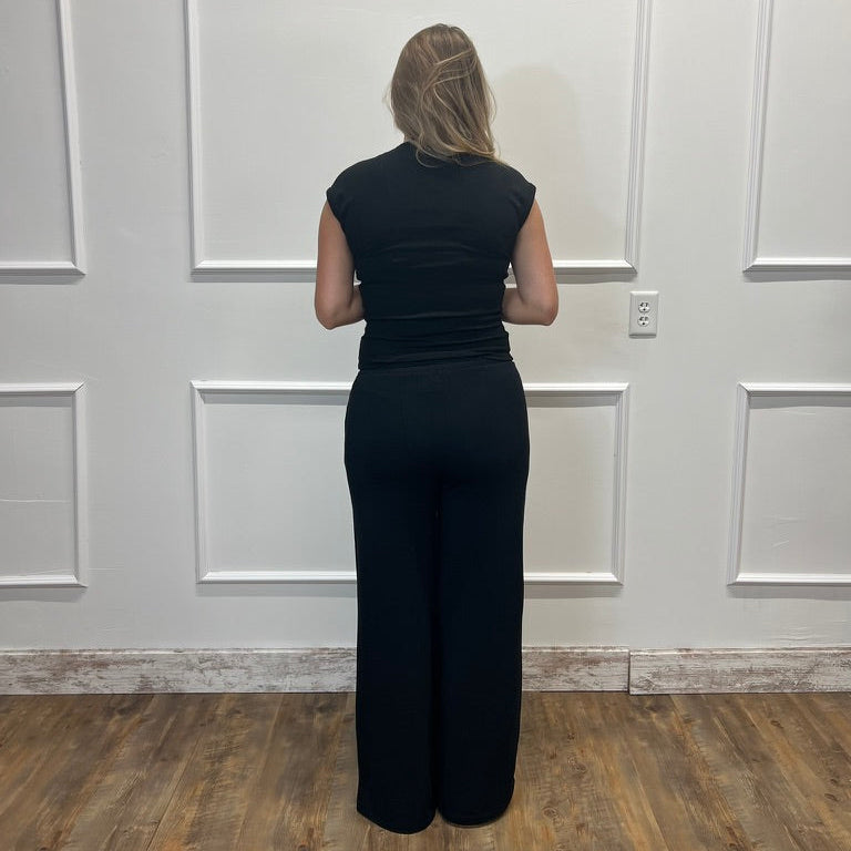 A woman viewed from behind wearing a black sleeveless jumpsuit with wide legs, standing in front of a white paneled wall on a wood floor.