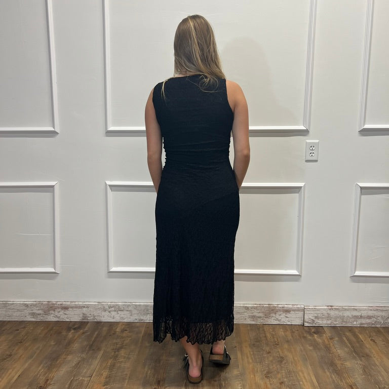 A woman viewed from behind wearing a sleeveless black maxi dress with a lace hem, standing in front of a white paneled wall on a wood floor.