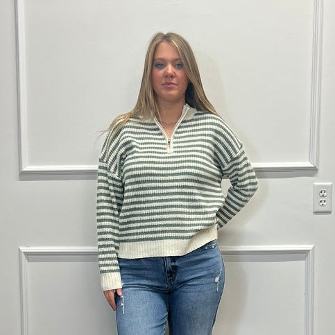 A woman stands in a room with white paneled walls and a wood floor, wearing a green and white striped half-zip sweater, blue jeans, and black slide sandals.