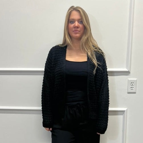 A woman stands indoors wearing a black textured knit cardigan over a black top and black pants, paired with black slide sandals. She is positioned in front of a white paneled wall on a wood-look floor.
