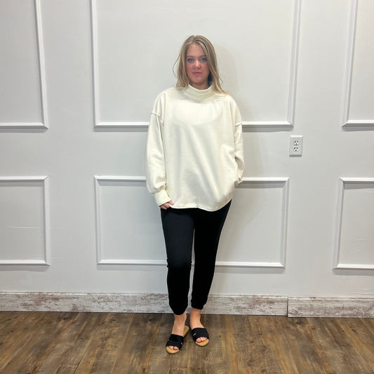 A woman stands in a room with white paneled walls and a wood floor, wearing an oversized cream mock-neck long-sleeve top, black pants, and black slide sandals.