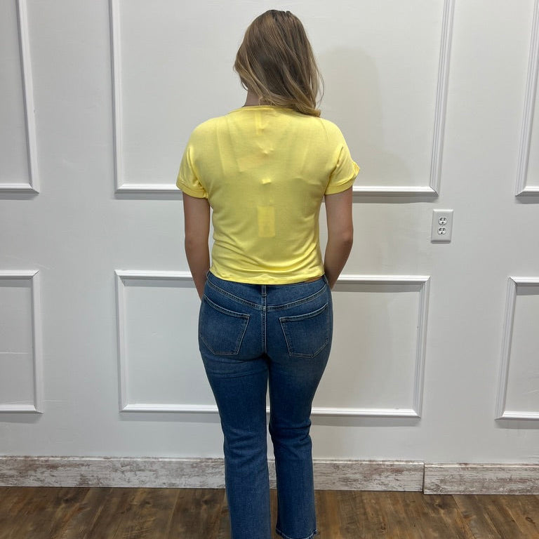 A woman viewed from behind wearing a bright yellow short-sleeve t-shirt and blue jeans, standing against a white paneled wall on a wood floor.