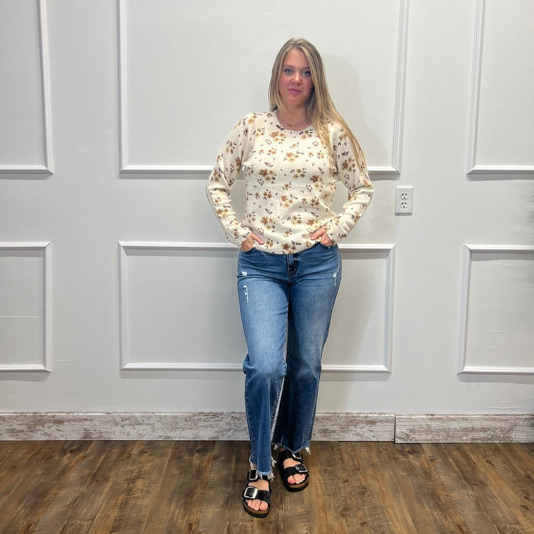 A woman stands in front of a white paneled wall wearing a cream long-sleeve top with a small yellow and green floral pattern, blue jeans, and black sandals.