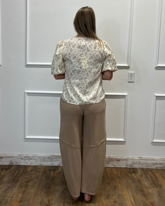 A woman stands with her back to the camera wearing a cream-colored short-sleeve blouse with a small floral pattern and beige wide-leg pleated pants, set against a white paneled wall and wood floor.