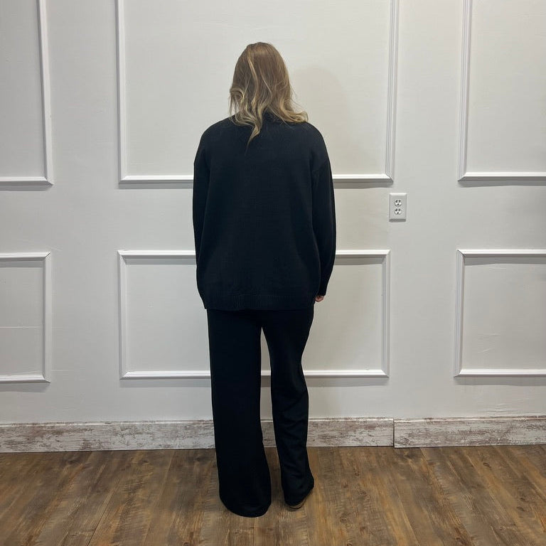 A woman stands with her back to the camera wearing a black long-sleeve top and black wide-leg pants, standing in front of a white paneled wall on a wood floor.