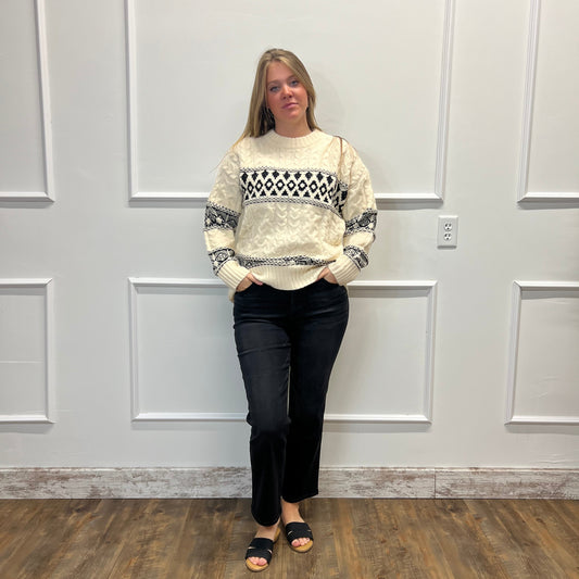 A woman stands in a room with white paneled walls and a wood floor, wearing a cream knit sweater with a black geometric pattern across the chest and sleeves, paired with black pants and black slide sandals.