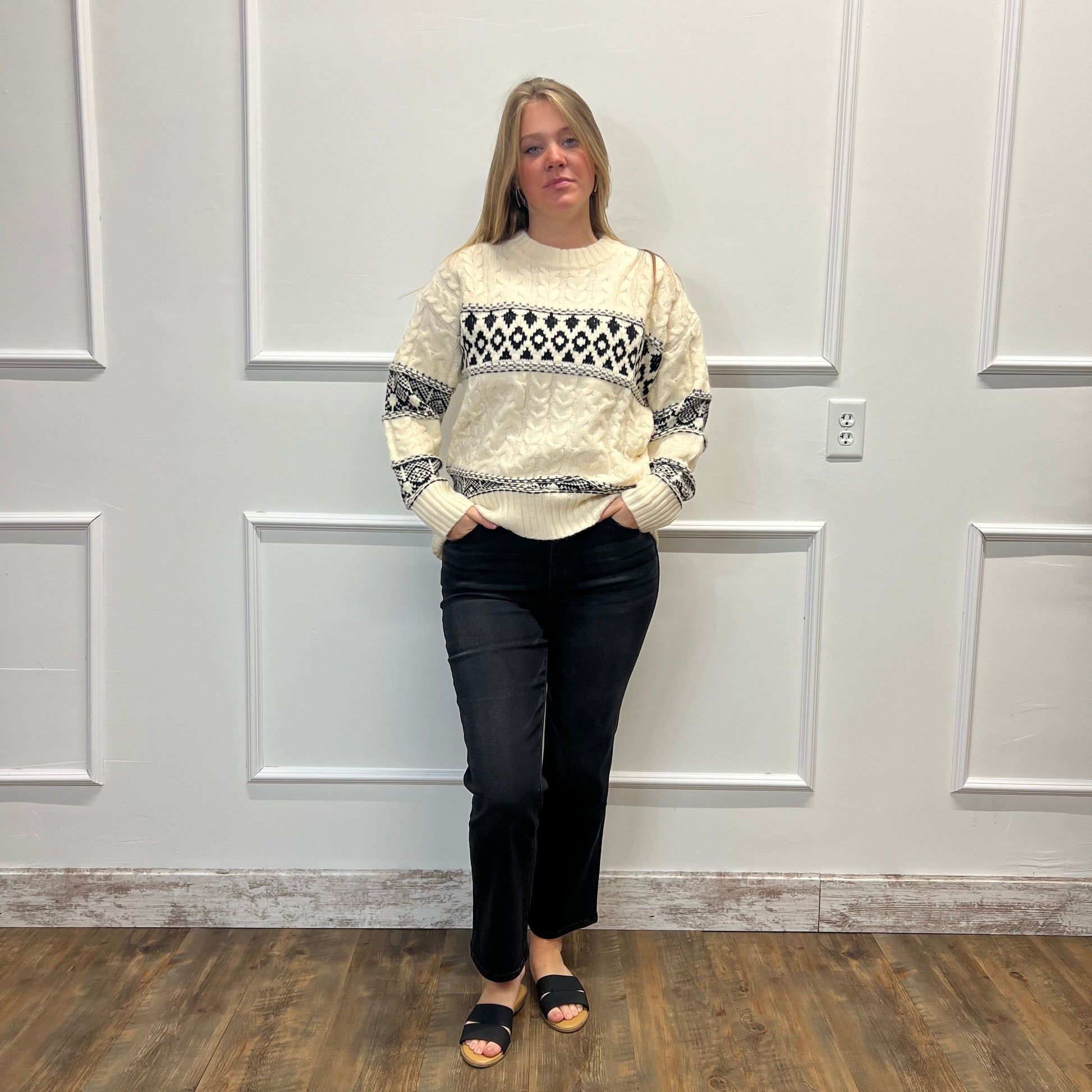 A woman stands in a room with white paneled walls and a wood floor, wearing a cream knit sweater with a black geometric pattern across the chest and sleeves, paired with black pants and black slide sandals.