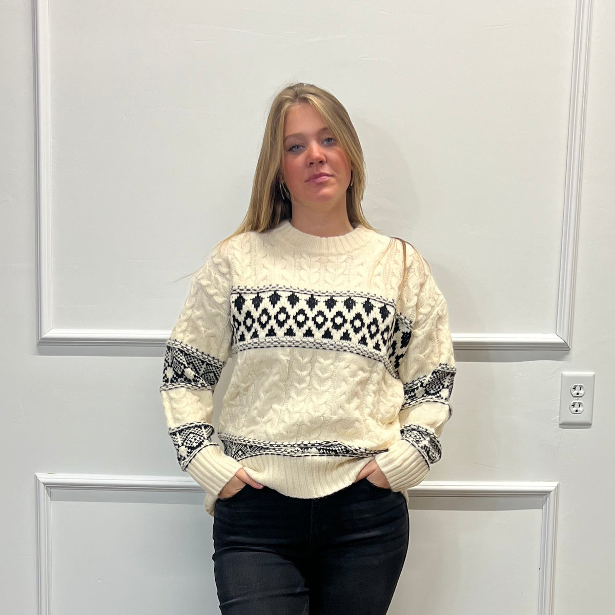 A woman stands in a room with white paneled walls and a wood floor, wearing a cream knit sweater with a black geometric pattern across the chest and sleeves, paired with black pants and black slide sandals.