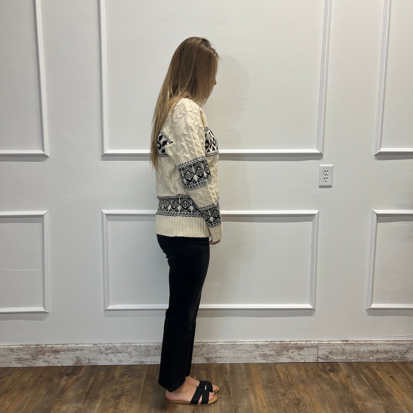 A woman stands in profile wearing a cream cable-knit sweater with black and gray Nordic-style patterns, paired with black pants and black slide sandals, set against a white paneled wall and wood-look floor.